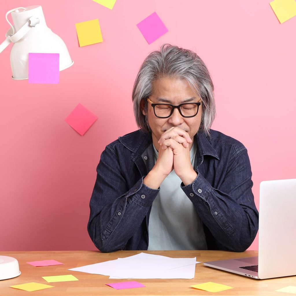 A senior Asian man is seated and praying at his work desk. The desk has a prayer list and lots of post-it notes on the desk, the lamp, the laptop, and the pink wall behind him.