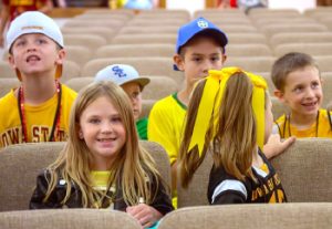 six middler- or junior-age kids (half girls, half boys) dressed in yellow or green casual clothes sit in a church auditorium looking happy